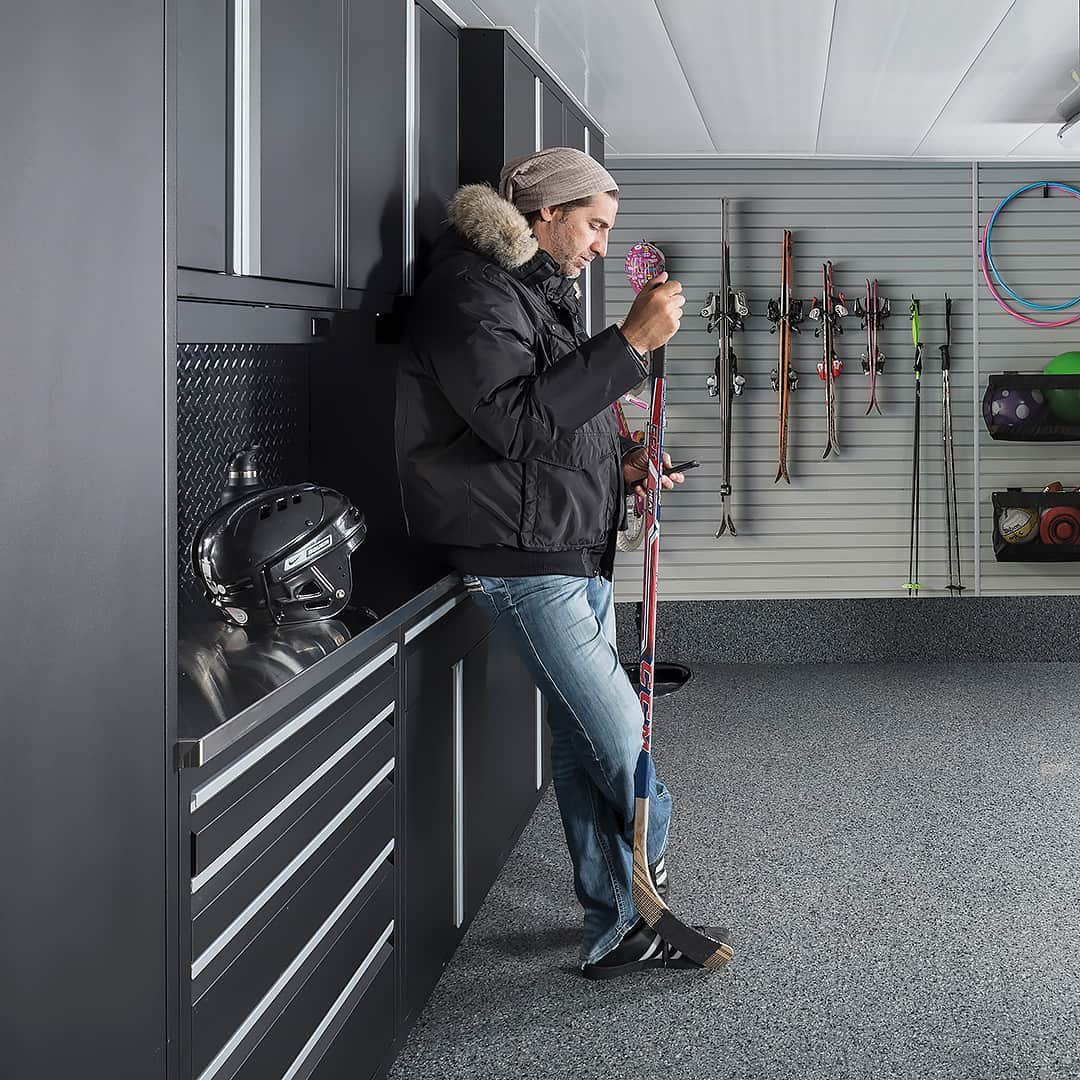 Man in winter coat standing in garage leaning on hockey stick.