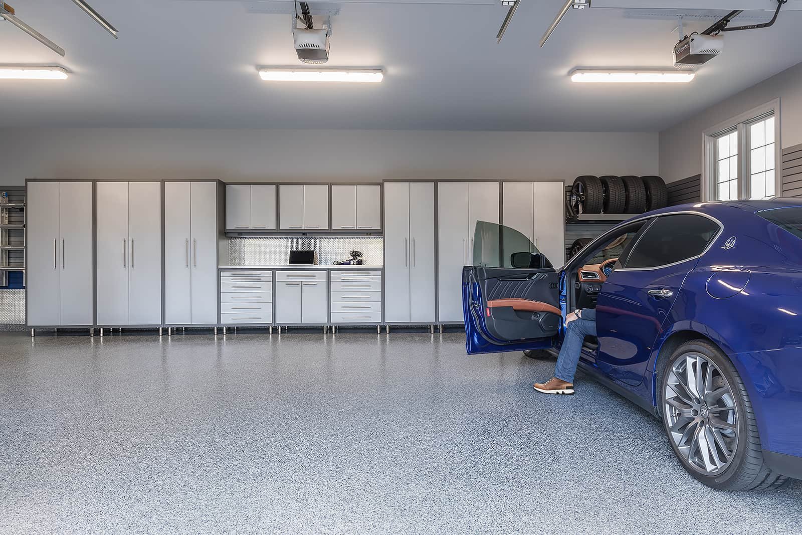 Man in winter coat stepping out of blue vehicle in large 3-car garage with garage cabinets.