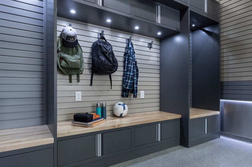 Garage storage area with dark cabinetry, wood bench, slatwall hooks holding backpacks and clothing, and a soccer ball on the bench.