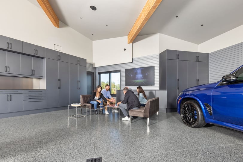 Finished garage with grey floor, wall-to-wall cabinetry, seating area, television, and blue vehicle parked alongside the living space.