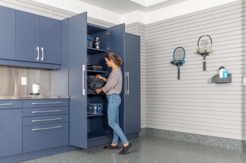 Woman placing a cooler inside tall blue garage cabinets with adjustable shelving beside a wall-mounted slatwall system.
