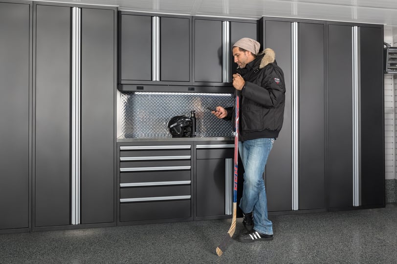 Man holding a hockey stick in front of dark grey garage cabinets with a helmet and water bottle on the work surface.